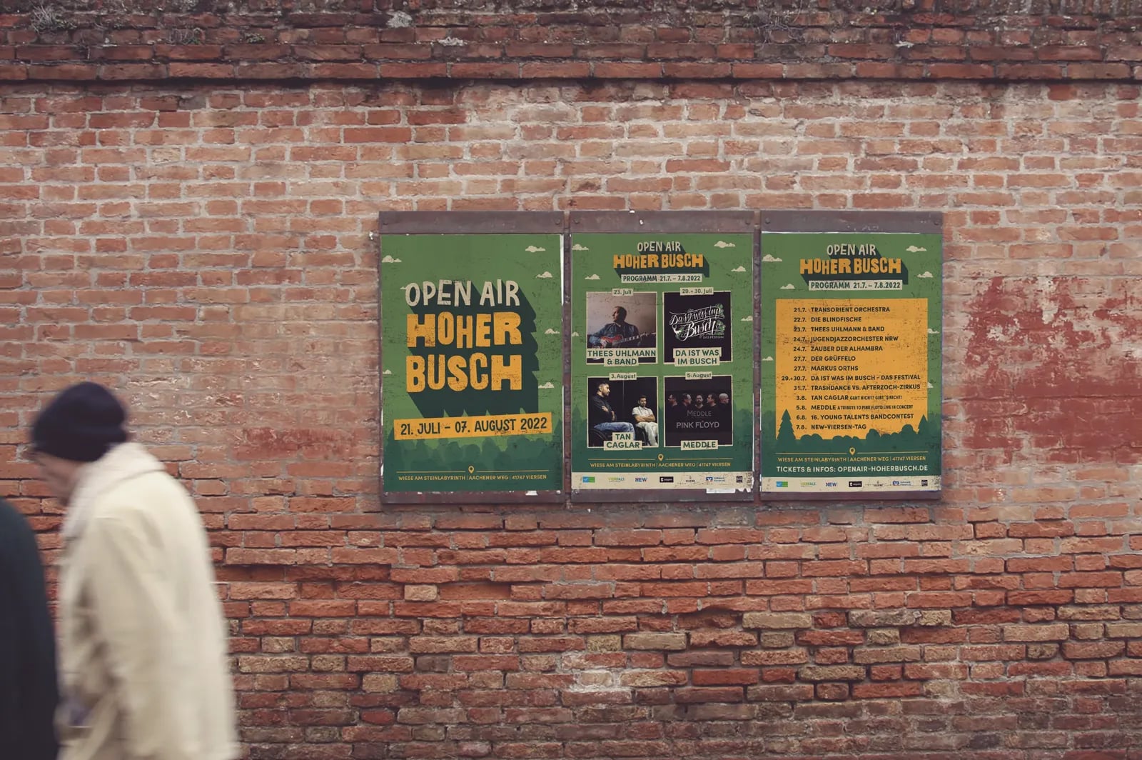 Plakate des Open Air Hoher Busch in einem Schaukasten an einer Backsteinwand, an der ein Passant vorbeigeht.